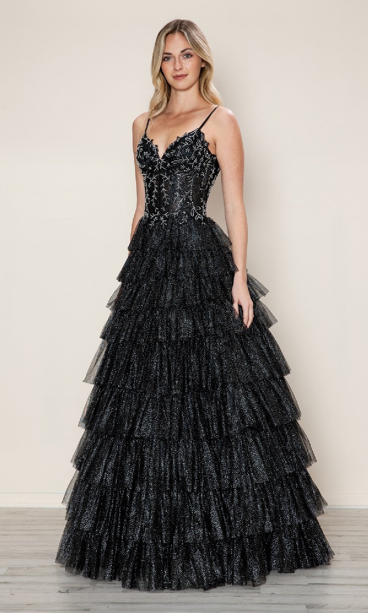 A woman stands against a plain background wearing the Long Prom Dress: Poly USA 9608, featuring a ruffled tiered tulle skirt and floral embroidery on the lace corset bodice.